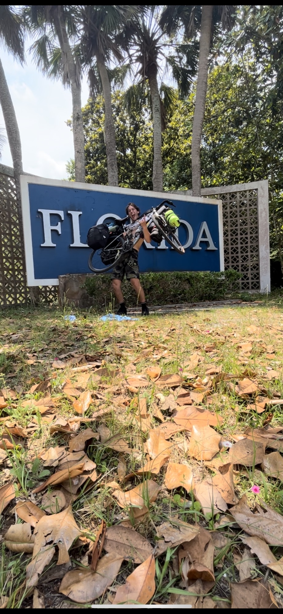 Pat hoisting his fully loaded bike overhead in front of the Florida welcome sign, 1,800 miles from home